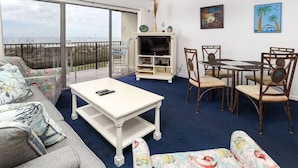 A living room with a sofa, coffee table, TV stand, and dining area with chairs. - Emerald Towers West 1003 - Sandy Toes (Fort Walton Beach)