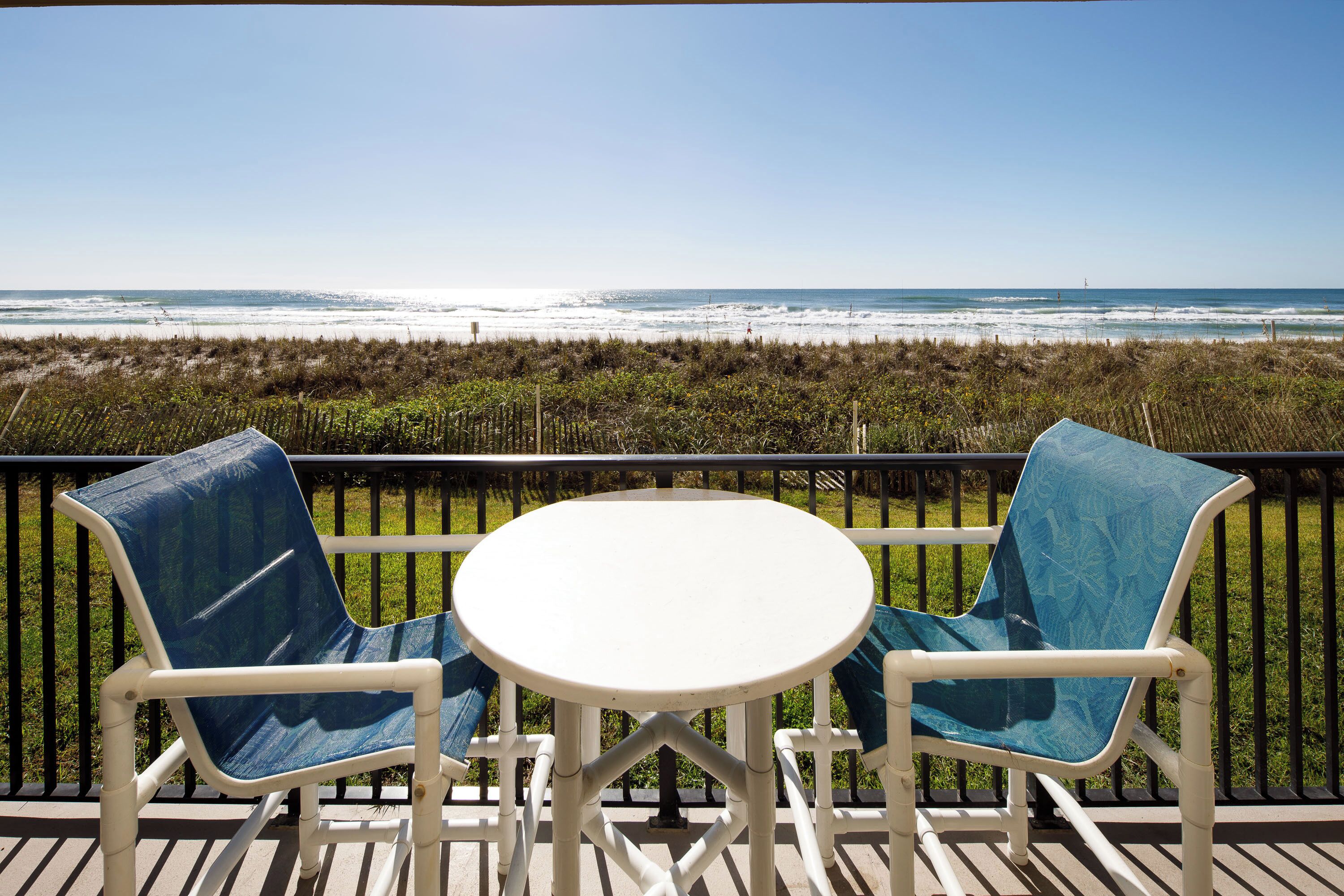 A balcony with two blue chairs and a white table, overlooking a beach with ocean and dunes.