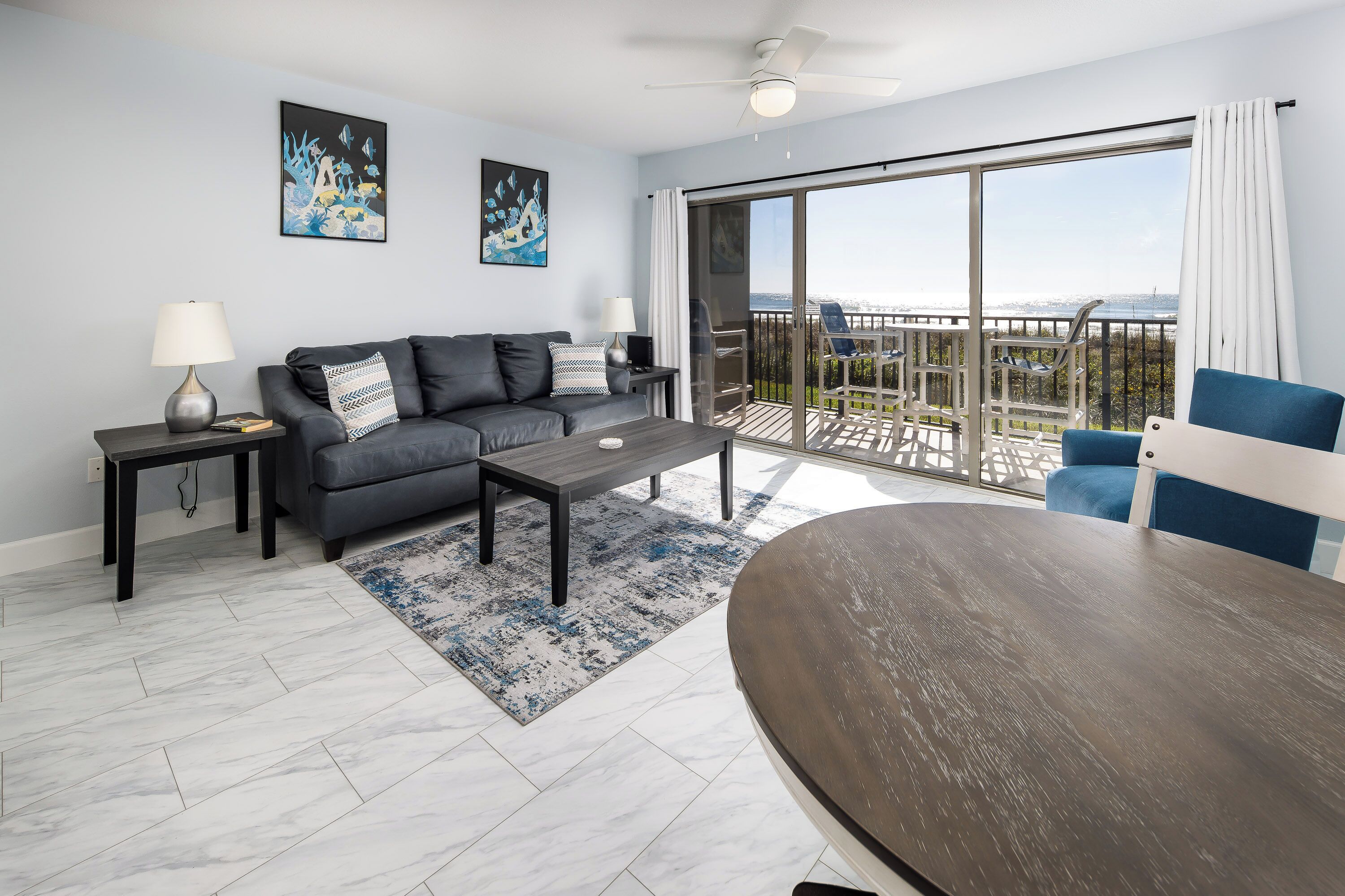 Un salon moderne avec un canapé gris, une table basse et un fauteuil bleu. Il y a un balcon avec vue sur les montagnes.