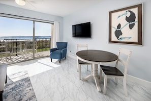 A modern room with a balcony, a flat-screen TV, a framed artwork, and a dining table with chairs. - Emerald Towers West 1003 - Sandy Toes (Fort Walton Beach)
