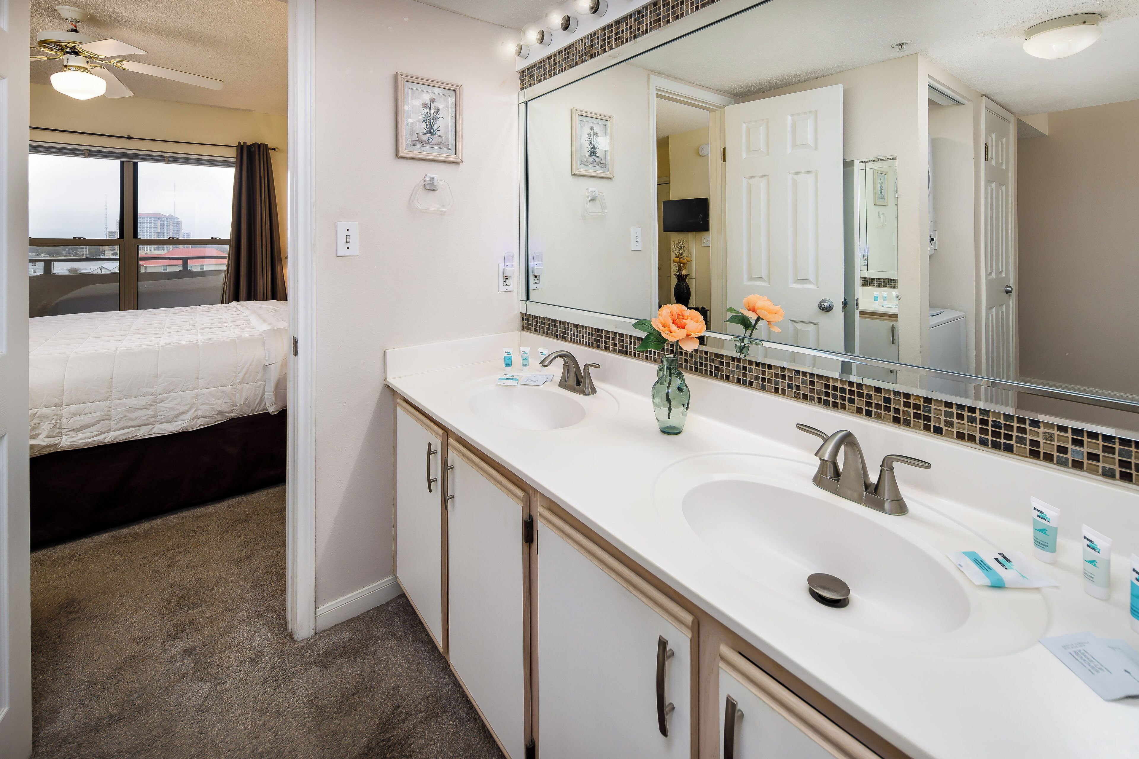 A hotel bathroom with a large mirror, two sinks, and a bed in the adjacent room.