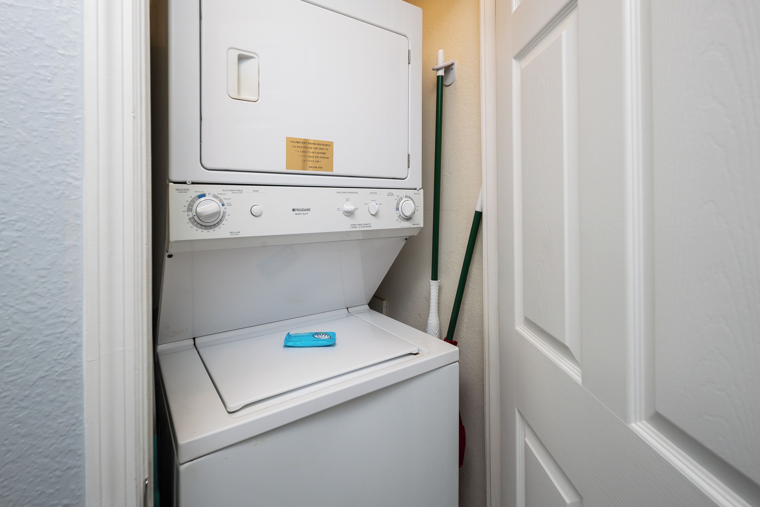 A compact laundry room with a front-loading washer and dryer, a notice on the dryer, and a green mop leaning against the wall.