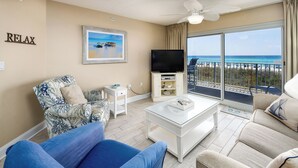 Un salon avec une grande télévision, une table basse et un canapé. Il y a un balcon avec vue sur la plage.