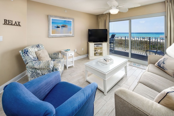 A living room with a large window overlooking a beach, a flat-screen TV, and a ceiling fan. - Summerlin 103 - Shell Seekers Retreat (Fort Walton Beach)