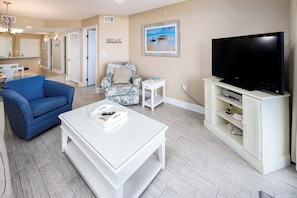 A living room with a blue sofa, a white coffee table, a patterned armchair, a TV on a white stand, and a painting of a beach scene. - Summerlin 103 - Shell Seekers Retreat (Fort Walton Beach)