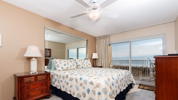 Une chambre avec un grand lit, une commode, un ventilateur de plafond, une grande fenêtre donnant sur un beau paysage et un balcon.