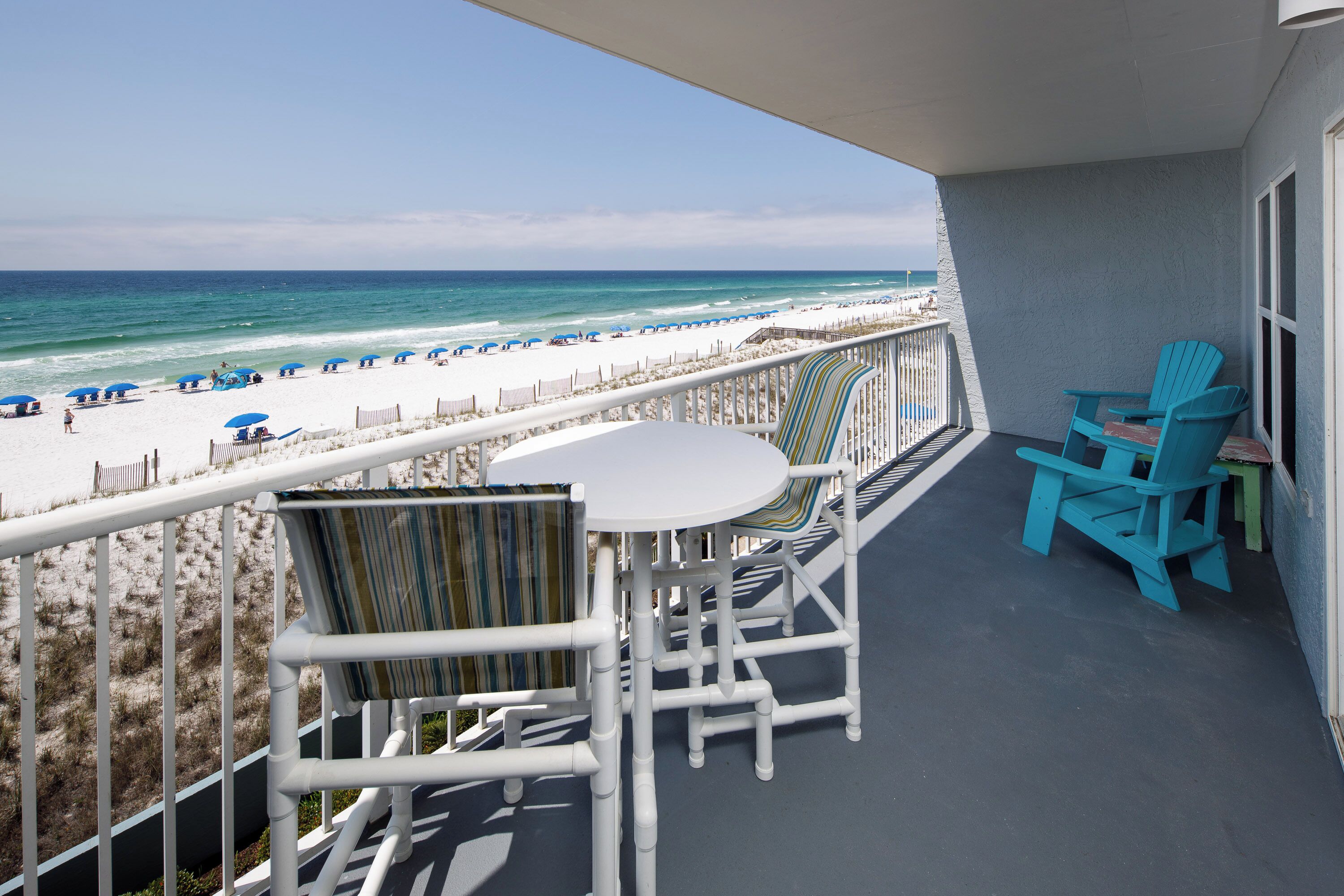 Un balcon avec vue sur la plage, des chaises blanches, une table et un fauteuil Adirondack bleu.