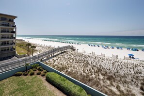 A beach view with a wooden boardwalk leading to the shore, a sandy beach with umbrellas, and a building with multiple floors. - Islander 4002 - Whispering Waves (Fort Walton Beach)