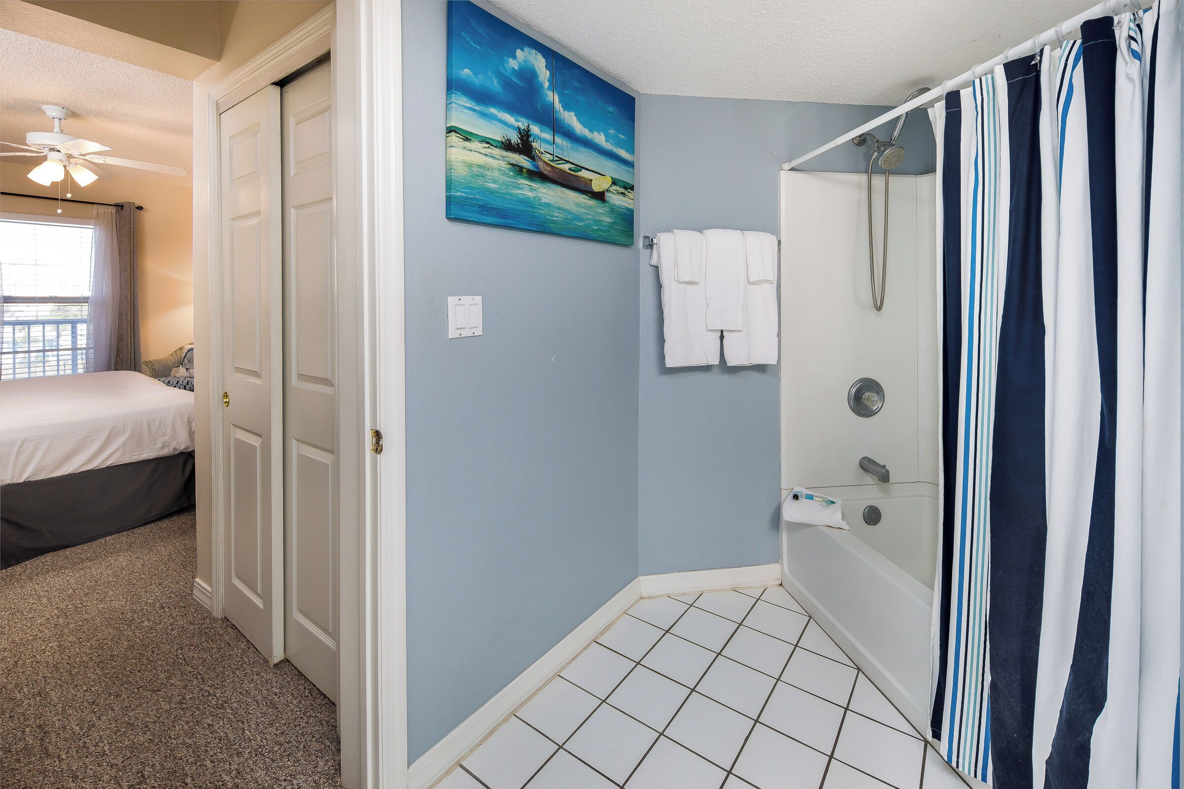 A bathroom with a shower, a painting on the wall, and a bed in the adjacent room.