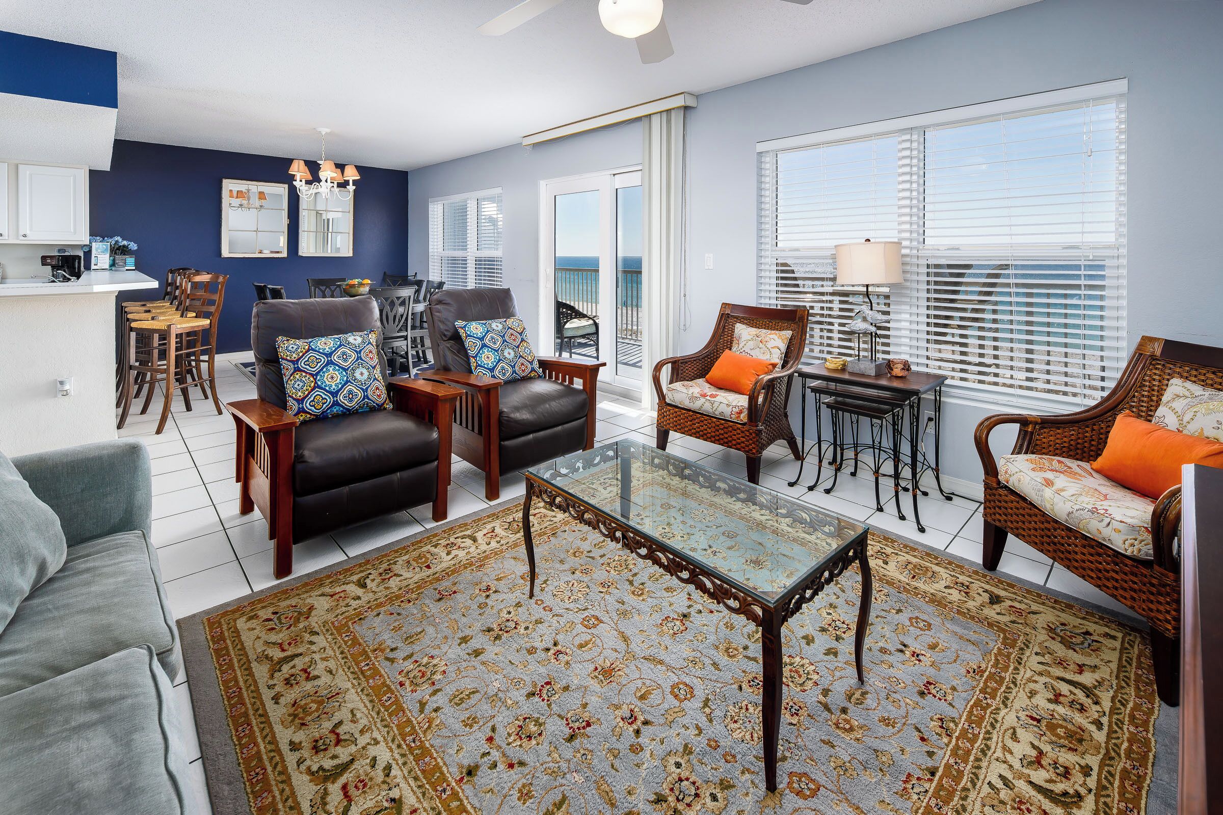 A spacious living room with a glass coffee table, a sofa, and two armchairs. There is a kitchen area with a bar and a dining table.