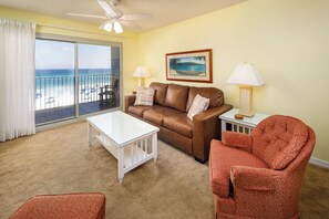 Ein Wohnzimmer mit einem braunen Ledersofa, einem weißen Couchtisch und einem roten, gemusterten Sessel. Ein großes Fenster bietet Blick auf den Strand, und ein Deckenventilator ist angebracht.
