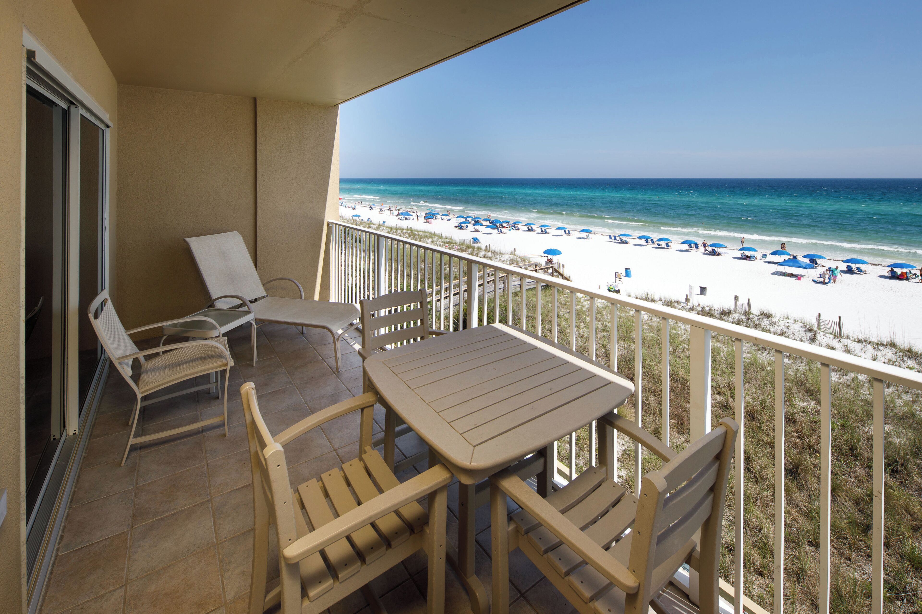 Een balkon met een tafel en stoelen, uitkijkend op een strand met parasols en mensen.