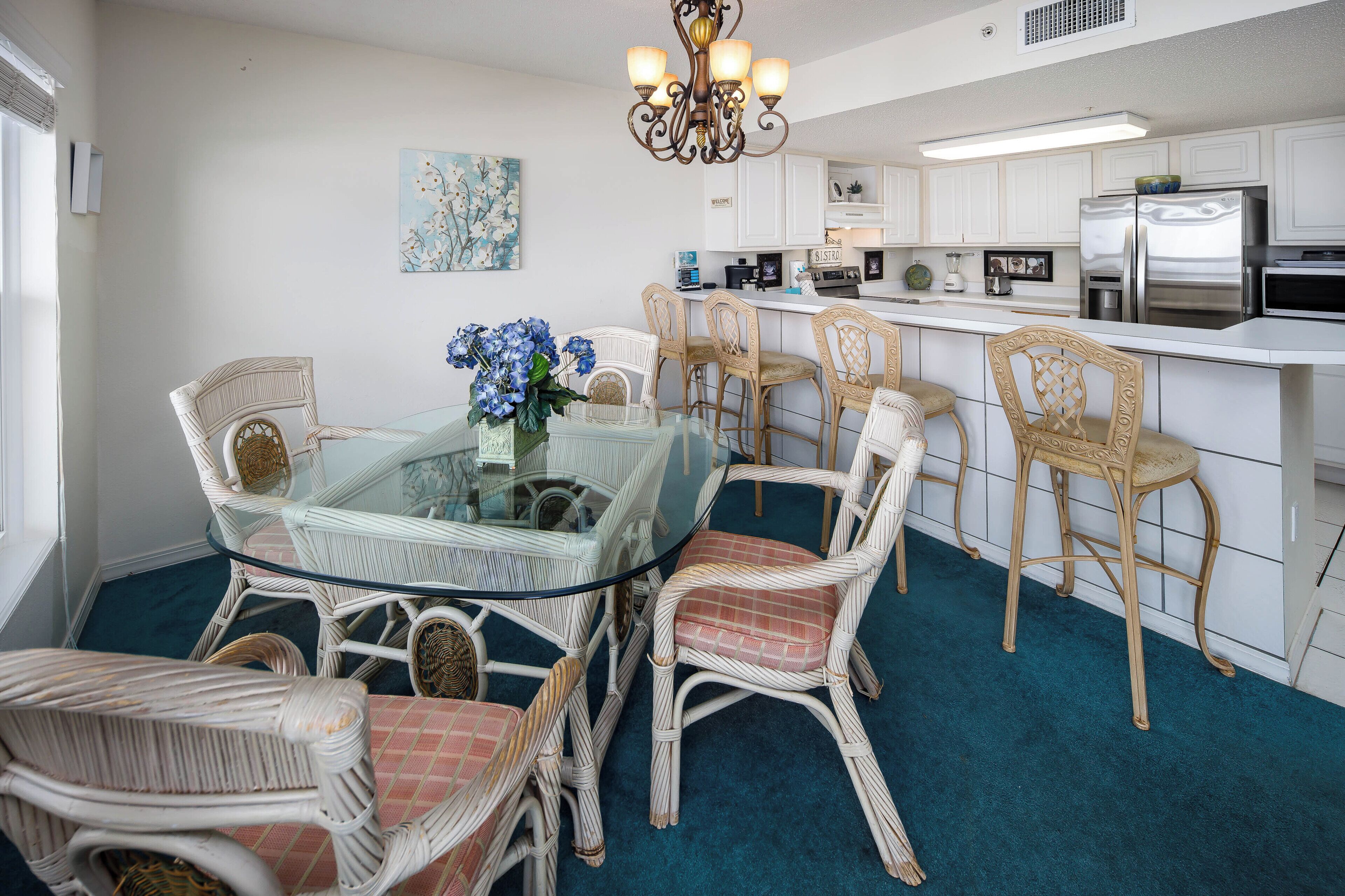 A dining area with a glass-top table surrounded by wicker chairs, a chandelier, and a blue carpet.