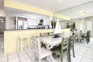 A modern kitchen with white cabinets, a central island with a sink, and a dining area with a glass table and chairs. - Islander 4011 by Brooks and Shorey Resorts (Fort Walton Beach)