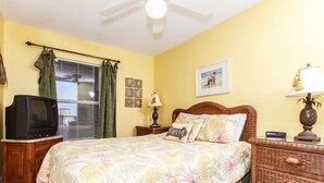 Un dormitorio con cama, televisor, ventilador de techo, ventana con cortinas y pared decorada con marcos.