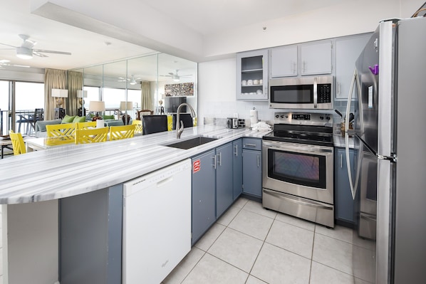 A modern kitchen with stainless steel appliances, a large island with a sink, and a dining area with yellow chairs. - Emerald Towers West 5006 - Coastal Vibe (Fort Walton Beach)