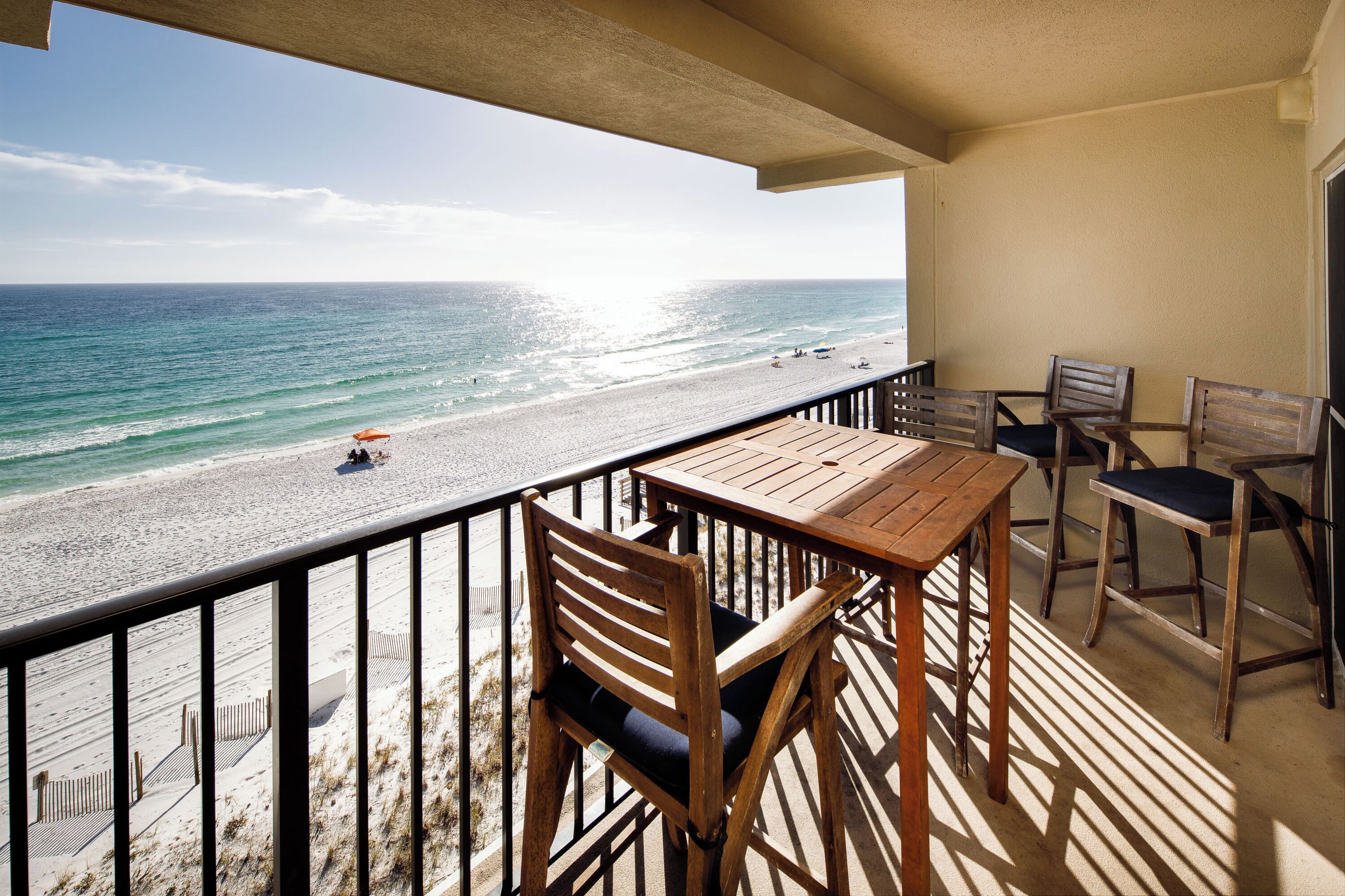 Un balcon avec une table et des chaises en bois, donnant sur une plage où se trouvent quelques personnes et un parasol.