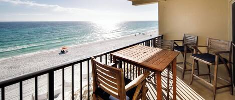 Un balcon avec une table et des chaises en bois, donnant sur une plage où se trouvent quelques personnes et un parasol.