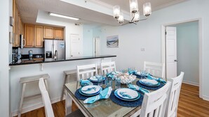 A dining area with a glass-top table set for six, white chairs, and a kitchen in the background. - Gulf Dunes 504 - Emerald Sea Oasis (Fort Walton Beach)