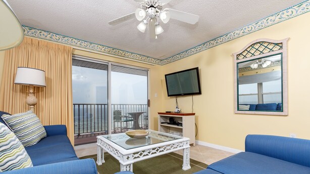 Een woonkamer met blauwe banken, een glazen salontafel, een plafondventilator, een televisie en een balkon met uitzicht.
