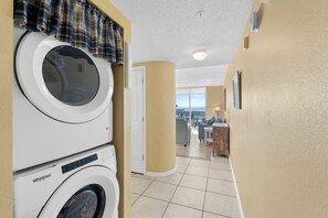 Uma lavanderia com máquinas de lavar e secar empilhadas, uma porta que dá para uma varanda com vista, e um pequeno espaço de estar com mesa e cadeiras.