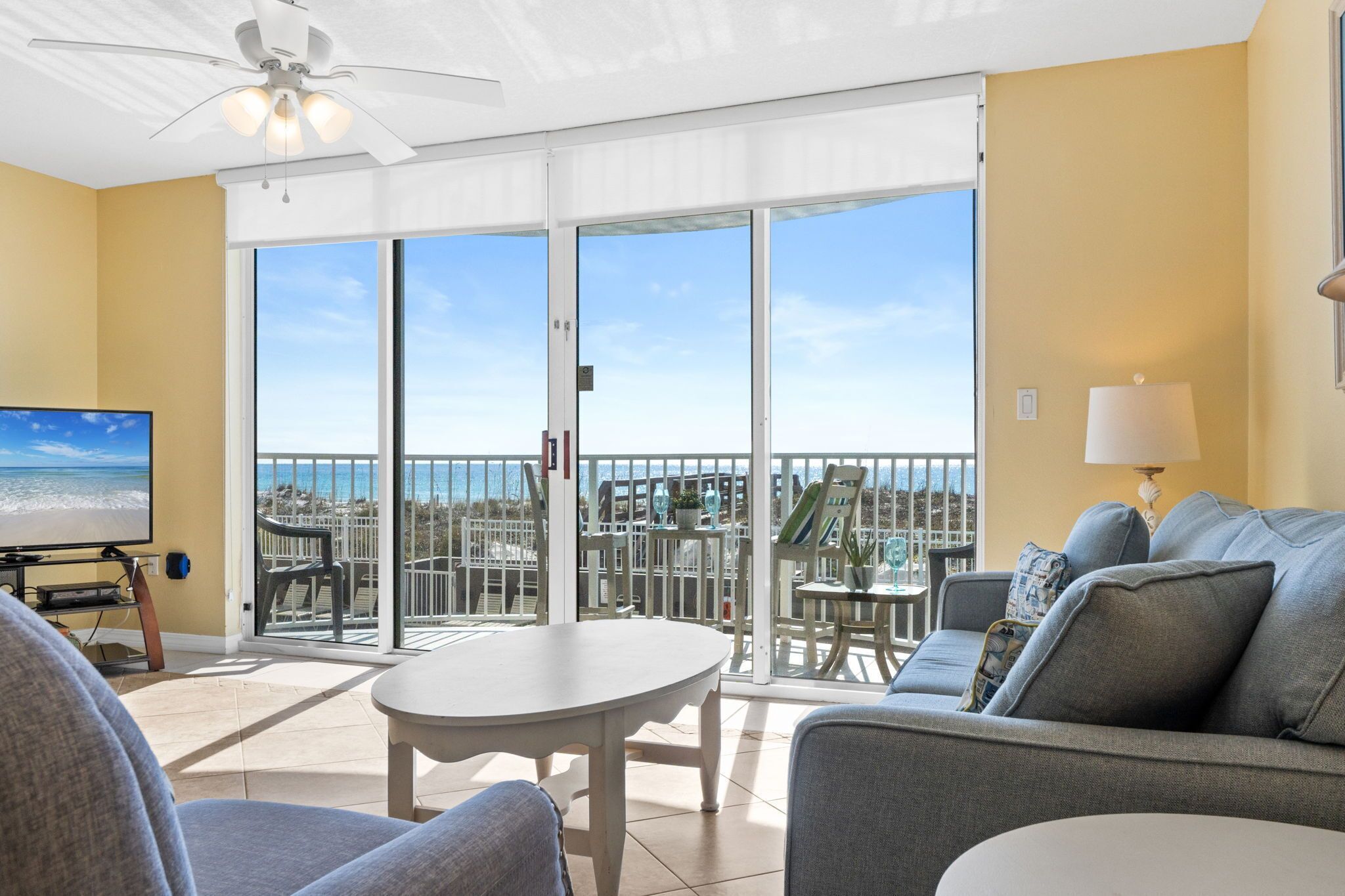 A balcony with a view of the ocean, a ceiling fan, a TV, and a sofa with a lamp.