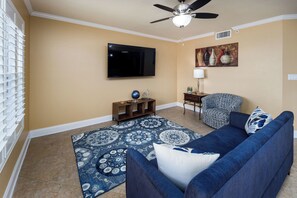 Ein Wohnzimmer mit blauem Sofa, einem gemusterten Teppich, einem Flachbildfernseher, einem Deckenventilator und einem Gemälde an der Wand.