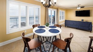 A dining area with a round table, six wooden chairs, a chandelier, and a view of the ocean through large windows. - Islander 4006 - Blue Waters and Beyond (Fort Walton Beach)