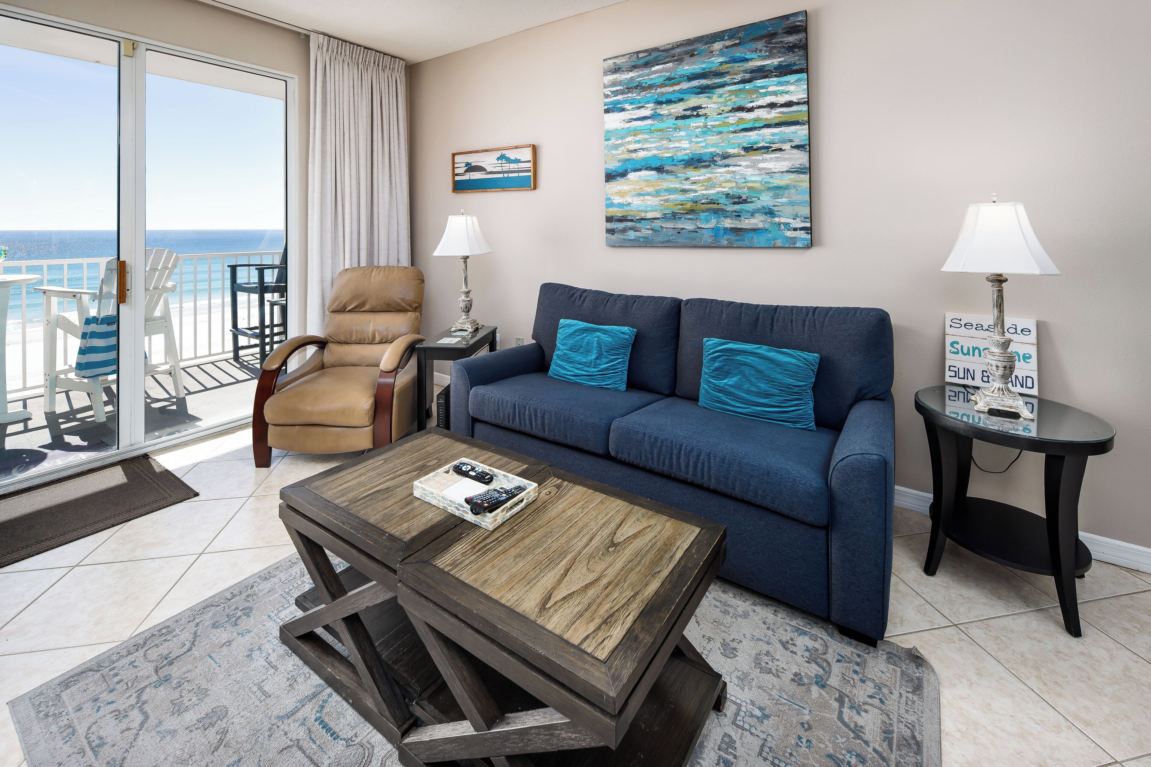 A living room with a blue sofa, a wooden coffee table, and a view of the ocean through a sliding glass door.