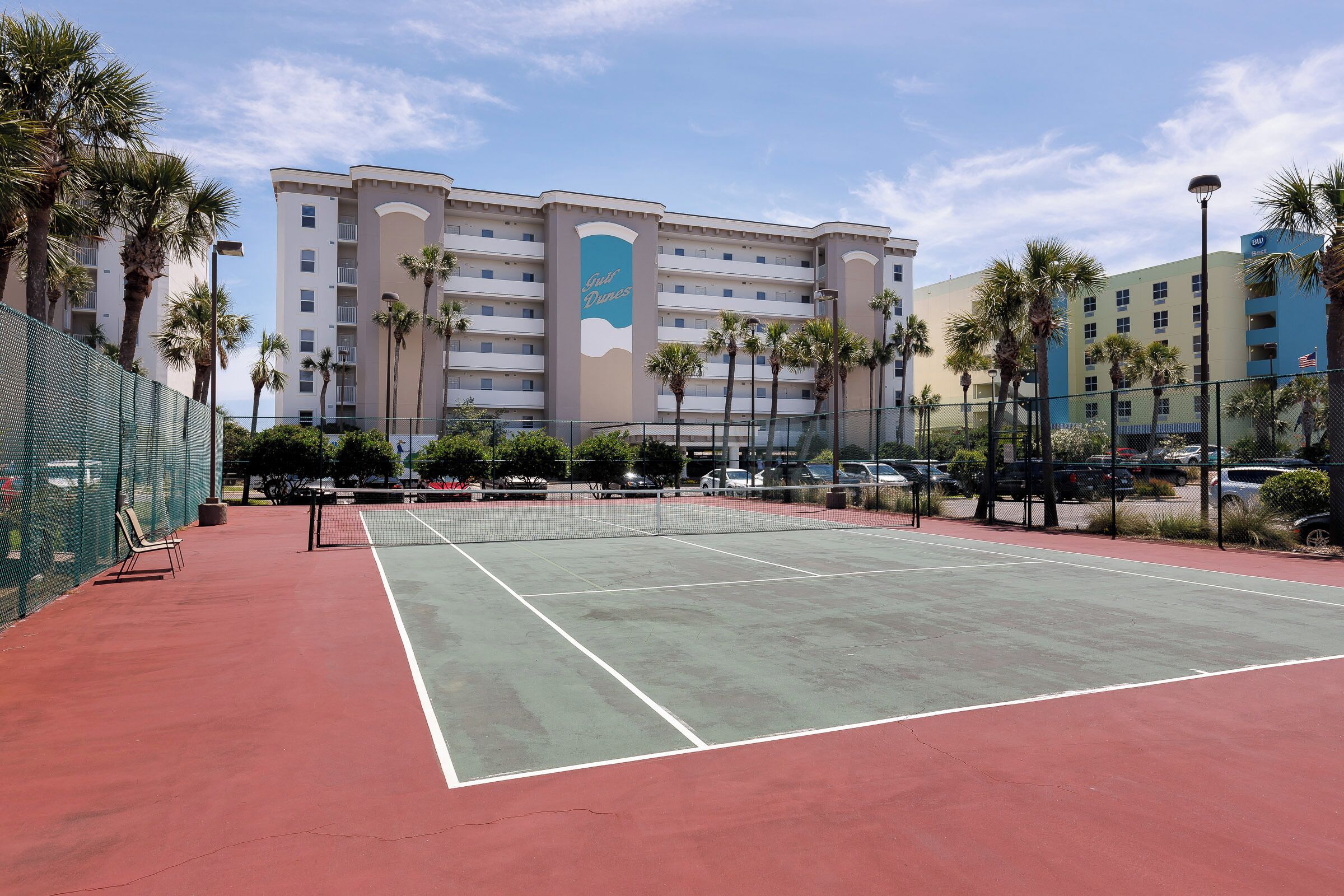Una cancha de tenis con red, rodeada de palmeras y un edificio de hotel al fondo.