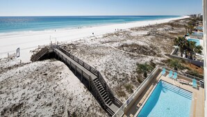 A beach view with a wooden boardwalk leading to a pool area with lounge chairs.