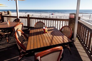 A wooden deck with tables and chairs, overlooking a beach with people and a sign warning about seagulls.