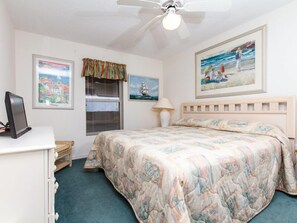 Une chambre à coucher comprenant un lit, un bureau, un ventilateur de plafond, une fenêtre avec des stores et des œuvres d’art accrochées aux murs.