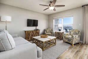 Un salon moderne doté d’un ventilateur de plafond, d’un téléviseur à écran plat, d’un canapé gris, de deux fauteuils à motifs et d’une table basse surmontée d’un panier en osier.