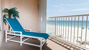Een balkon met een wit metalen hekwerk, een blauwe loungestoel, en uitzicht op een strand met helderblauw water en wit zand.