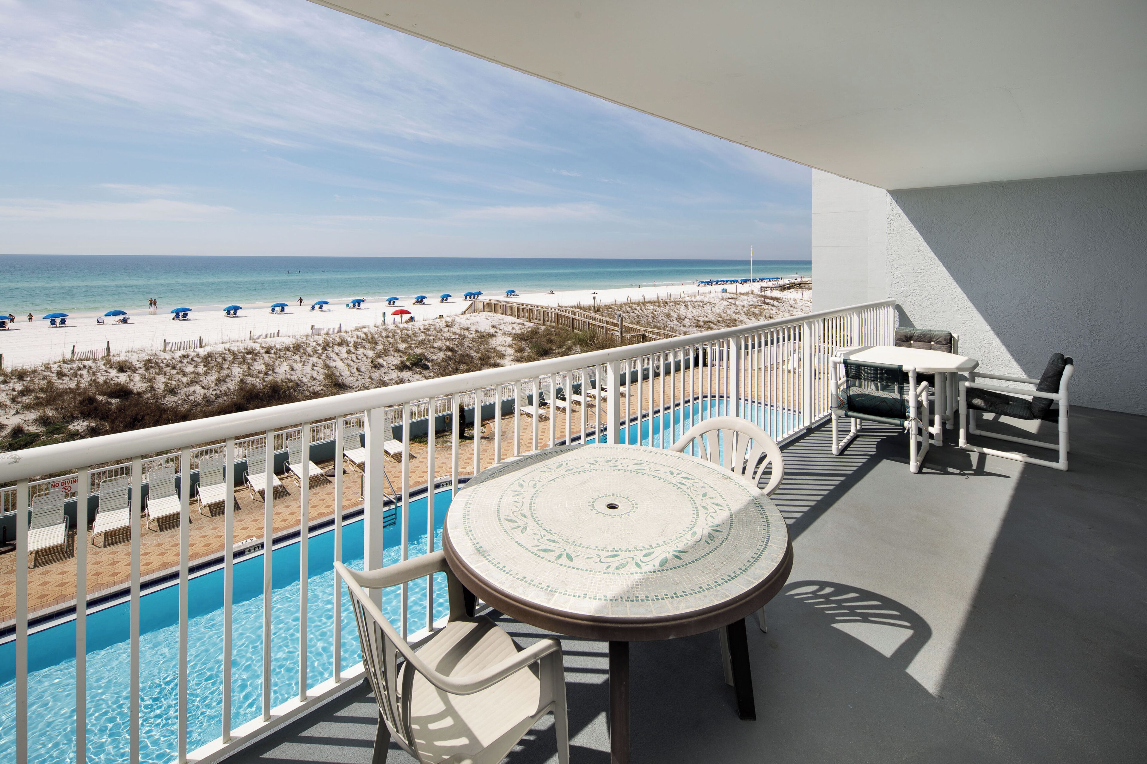 A balcony with a pool, white chairs, and a table. The balcony overlooks a beach with umbrellas and lounge chairs.