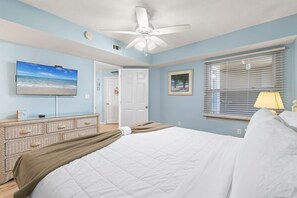 Ein Schlafzimmer mit einem Bett, einem Deckenventilator, einem Fernseher, einem Schminktisch und einem Fenster mit Jalousien sowie einer Tür.