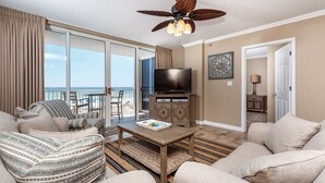 A spacious living room with a large television, a ceiling fan, and a view of the ocean through the sliding glass doors. - Island Princess 309 - Splash of Paradise (Fort Walton Beach)