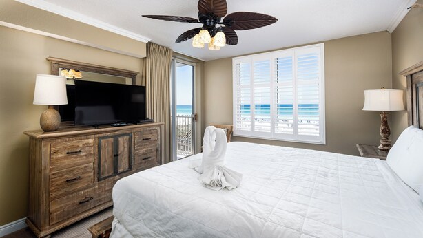 A hotel room with a large bed, a television on a wooden dresser, a ceiling fan, and a view of the beach through a window. - Island Princess 309 - Splash of Paradise (Fort Walton Beach)