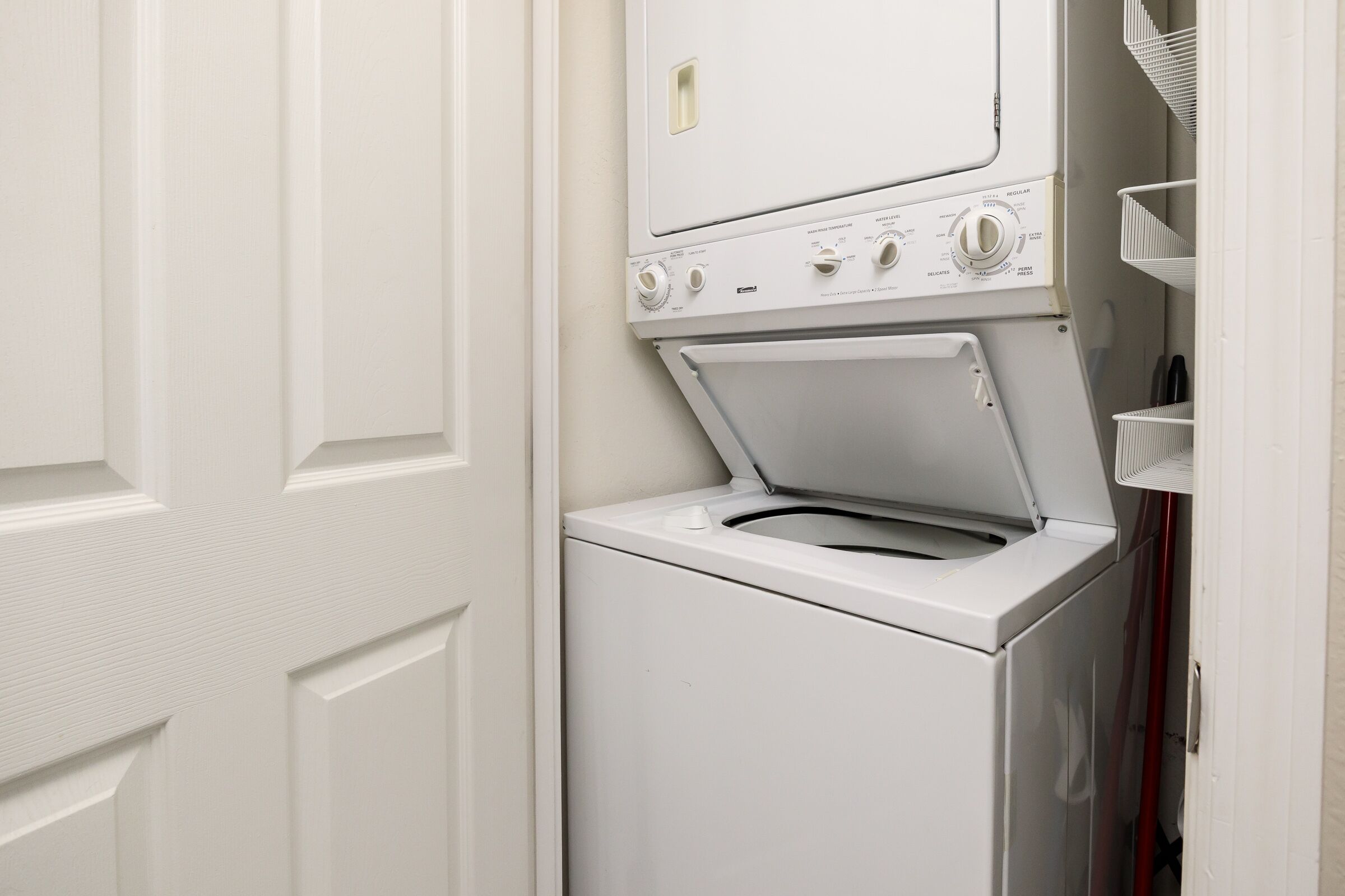 A compact laundry room with a front-loading washer and dryer, a white door, and a small shelving unit with baskets.