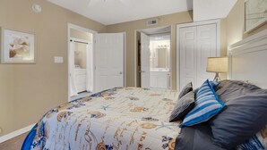 Un dormitorio con cama, ventilador de techo, baño con espejo y lavamanos, y una puerta que da a otra habitación.