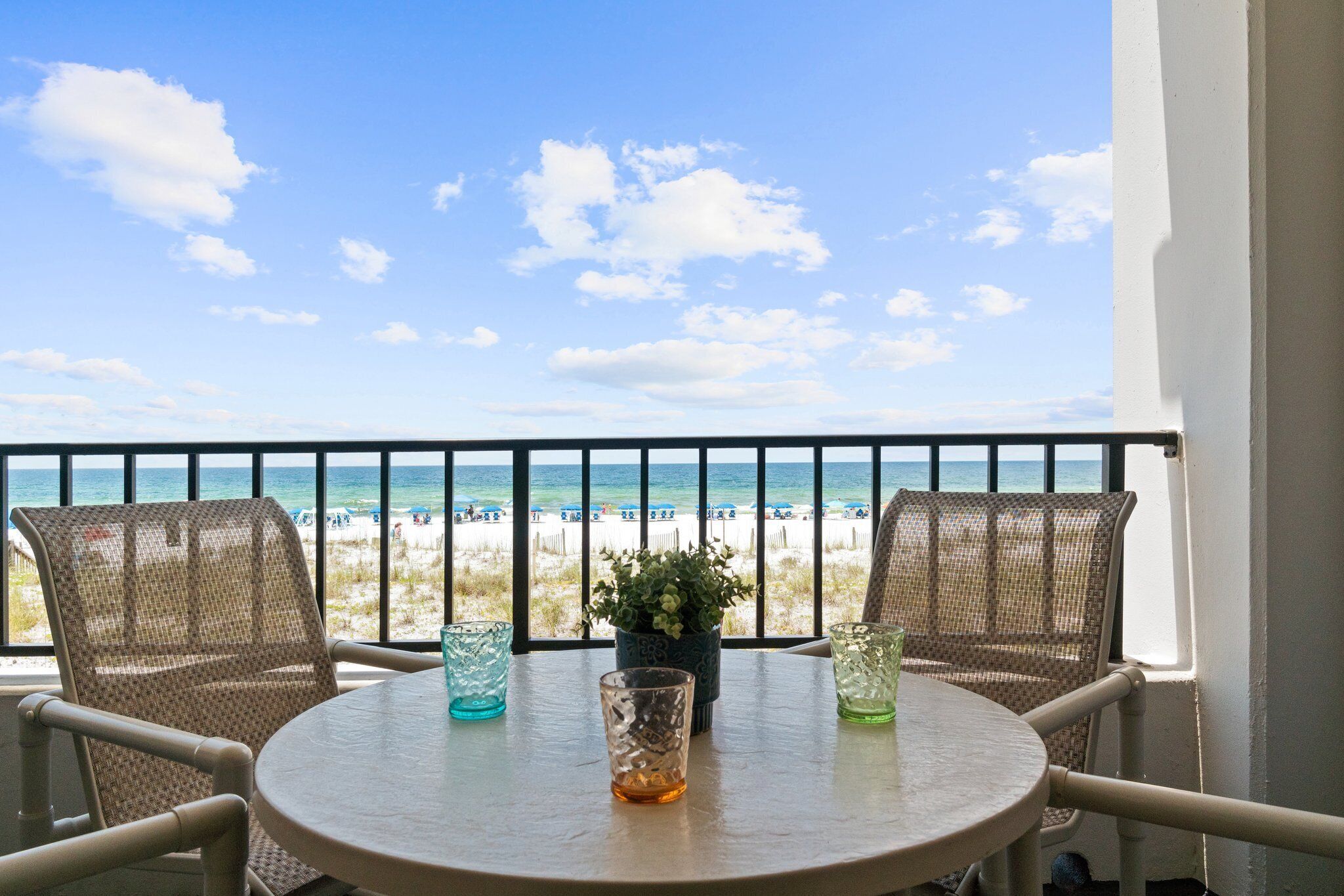 A balcony with two wicker chairs, a small table, and a potted plant. The view includes a beach and the ocean.
