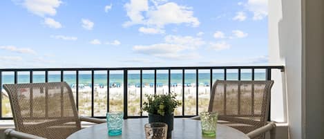A balcony with two wicker chairs, a small table, and a potted plant. The view includes a beach and the ocean.