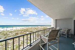 A balcony with wicker chairs and a table, overlooking a beach with umbrellas and a fence. - Surf Dweller 208 - Gulfview Oasis (Fort Walton Beach)