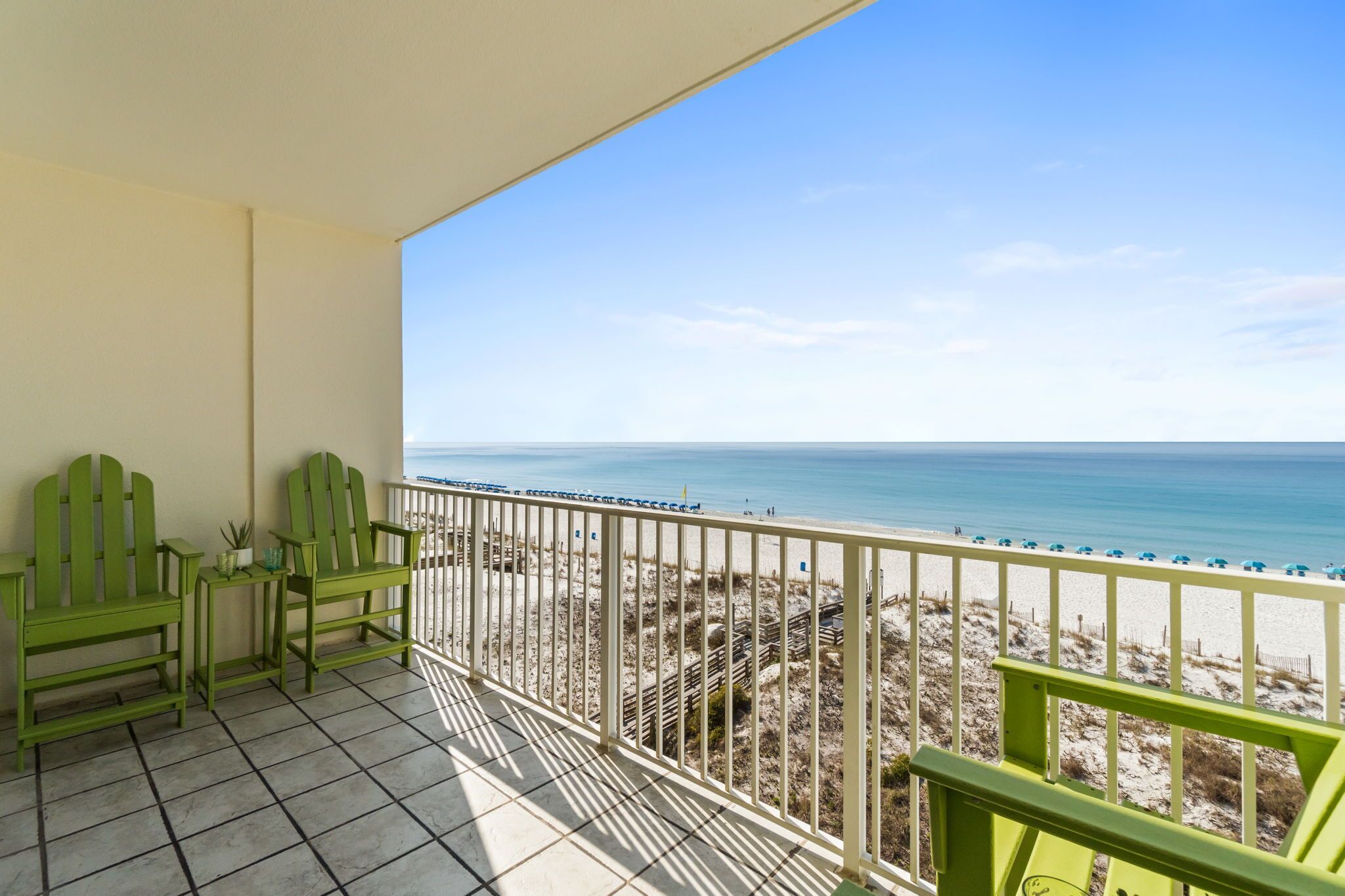 A balcony with a view of the beach, green chairs, and a tiled floor.