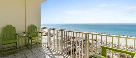 A balcony with a view of the beach, green chairs, and a tiled floor.