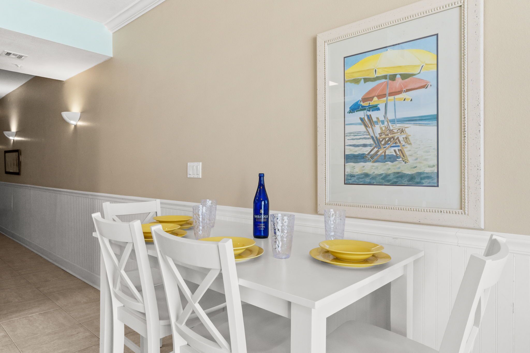 Un comedor con una mesa blanca dispuesta para cuatro personas, un cuadro enmarcado de una playa y una botella de SAKURA sobre la mesa.