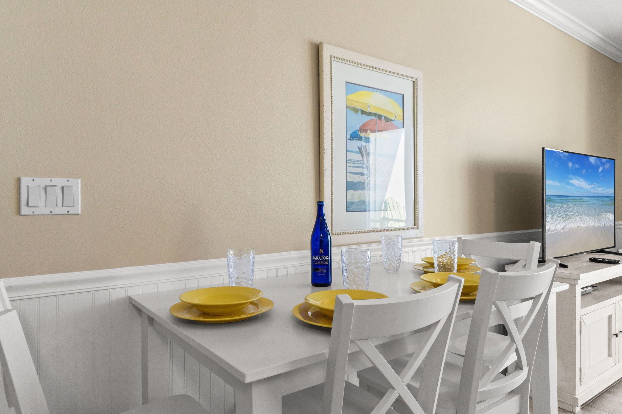 Un coin repas avec une table blanche dressée pour quatre personnes, des assiettes jaunes, des chaises blanches, un tableau encadré au mur, un téléviseur et une bouteille d’eau.