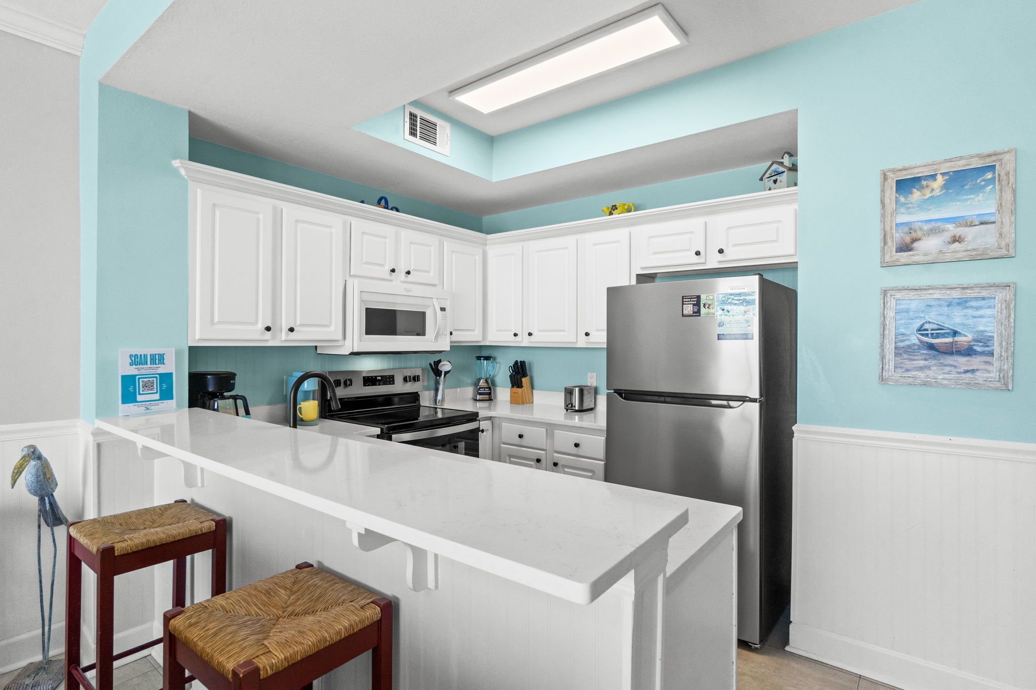 Cocina con armarios blancos, refrigerador de acero inoxidable, microondas y pileta. Hay dos sillas de mimbre y una pared azul con un cuadro.