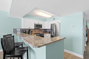 A modern kitchen with white cabinets, a granite countertop, stainless steel appliances, and a breakfast bar with two chairs. - Pelican Isle 508 - Sandcastle Shores (Fort Walton Beach)